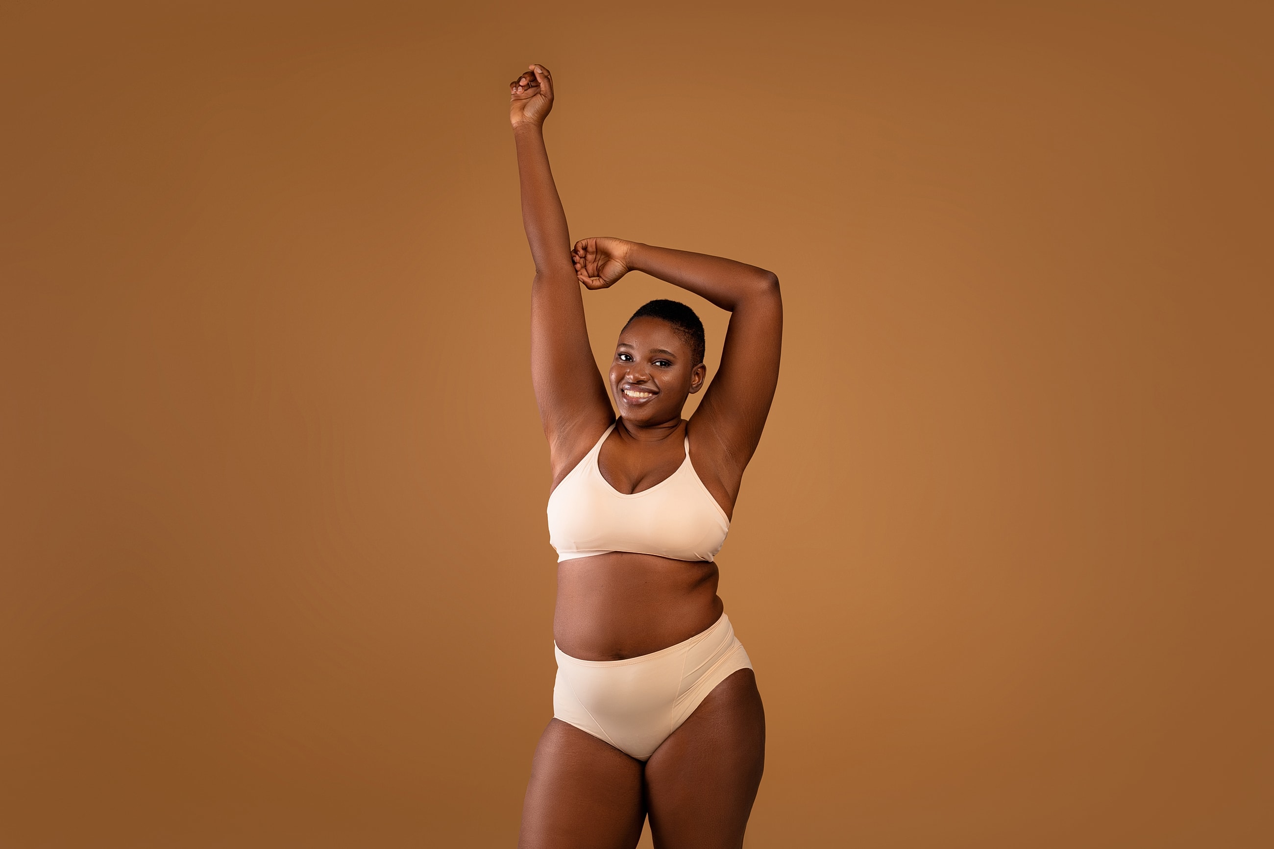 Smiling woman in beige lingerie with raised arms.